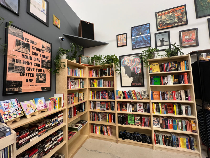Image depicting 5 bookshelves filled with books, VHS tapes, and shirts. Also shown are framed pictures, a speaker, and plants on top of the bookshelves