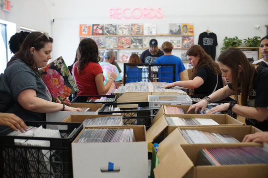 People sorting records in a store with boxes and shelves. Employees working at the front counter in the background.