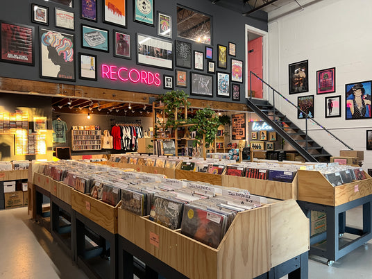 Rust & Wax record shop interior retail space. Many record bins on casters hold vinyl records in side of them. A gray gallery wall is above with a pink neon sign that says RECORDS and a bunch of concert posters and music themed artwork in frames.
