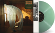 Image of Beck sitting on a chair in a room with wood paneling walls and an open window with curtains blowing inward. Beck Ride Lonesome sticker on top right corner of room. Vintage green vinyl record peeking out of the side of the sleeve.