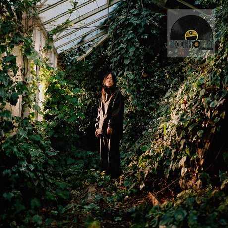 Deb Never standing amidst overgrown plants in a greenhouse with a record store day label logo in the corner.