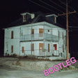 A nighttime photograph of a dilapidated white two-story house with boarded windows and a porch, set against a dark night sky with power lines and a utility pole. The word 'BOOTLEG' is written in large pink letters in the bottom right corner.