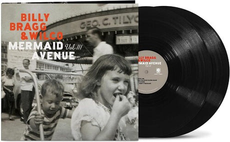 Vinyl album cover showing a child pushing another child on a hand cart in front of 3 other people and a large building (all in black & white) with orange text "Billy Bragg & Wilco" and white text "mermaid avenue vol. iii" with two black vinyl records sticking out the side of the sleeve.
