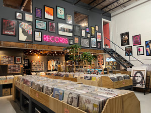 View from the front right corner of the shop, facing the main floor. Features 2 rows of record bins, as well as the staircase, gallery wall hanging above the floor, and the back walls with cassette, CD's, shirts, books and more.