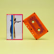 A retro-style cassette tape and its matching clear plastic case on a pale yellow background. The cassette is bright orange with purple text, and the case features a white cover with a black-and-white photo of a woman and a vertical rainbow stripe.