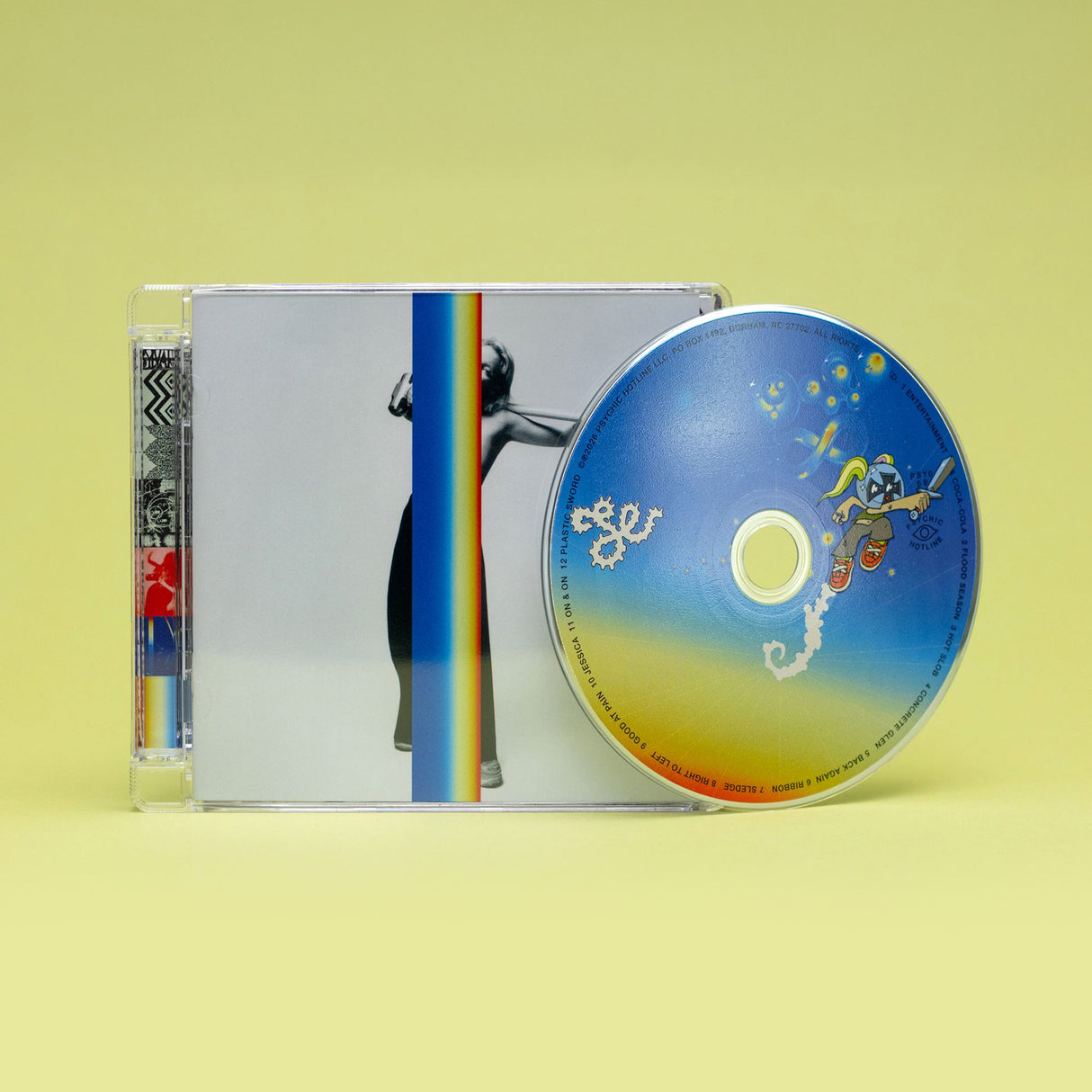A CD jewel case with a CD sitting on top of half of it, photographed on a pale yellow background. The album cover shows a dark figure against a white field with a vertical rainbow stripe, and the disc itself is blue with a yellow-to-orange gradient and small illustrated graphics.