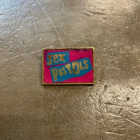 Sex Pistols Enamel Pin Front - Gold text on blue bars against pink backdrop