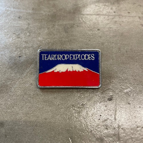 Teardrop Explodes Enamel Pin - Front with silver text against blue backdrop on top of red mountain